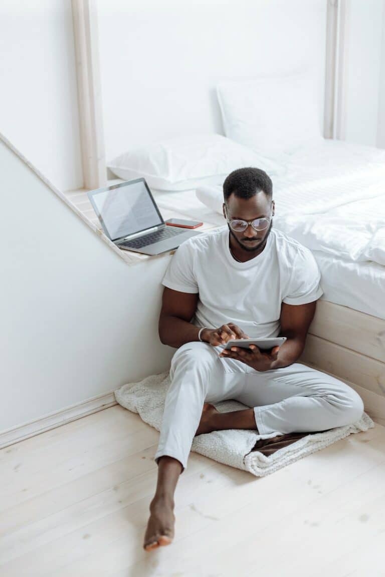 A Man Sitting on the White Blanket on the Floor while Using His Tablet