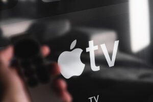 A close-up photo of a glossy screen displaying the Apple TV logo, with a reflection of a person's hand holding a remote control