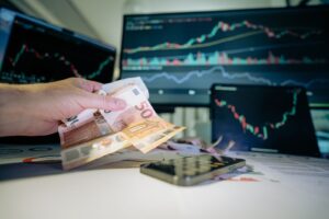 Business professional counting Euro notes with financial charts on screens, indicating investment strategy.